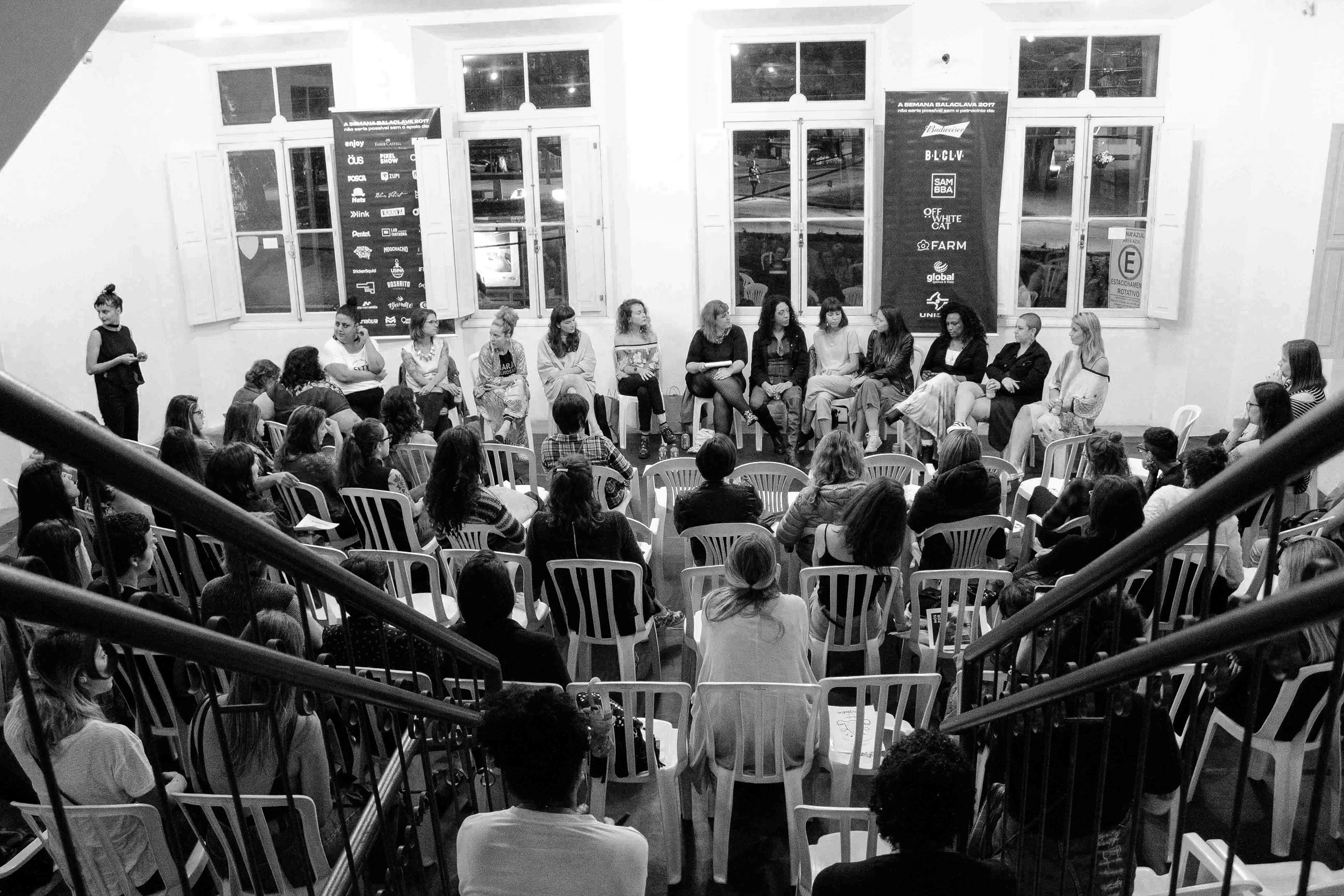 Attendees seated in a circle for a talk in a white gallery space, seen from a staircase.