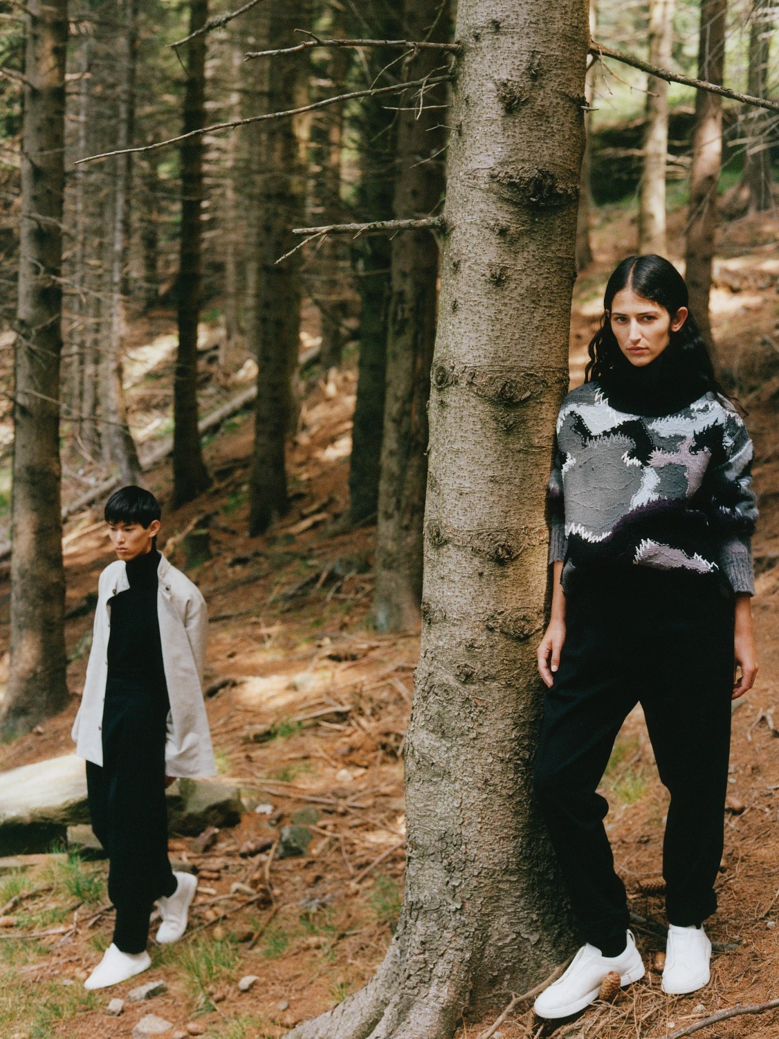 Two models in a conifer forest, one in a grey and black patterned cashmere turtleneck leaning against a tree trunk, the other in a pale coat further back.