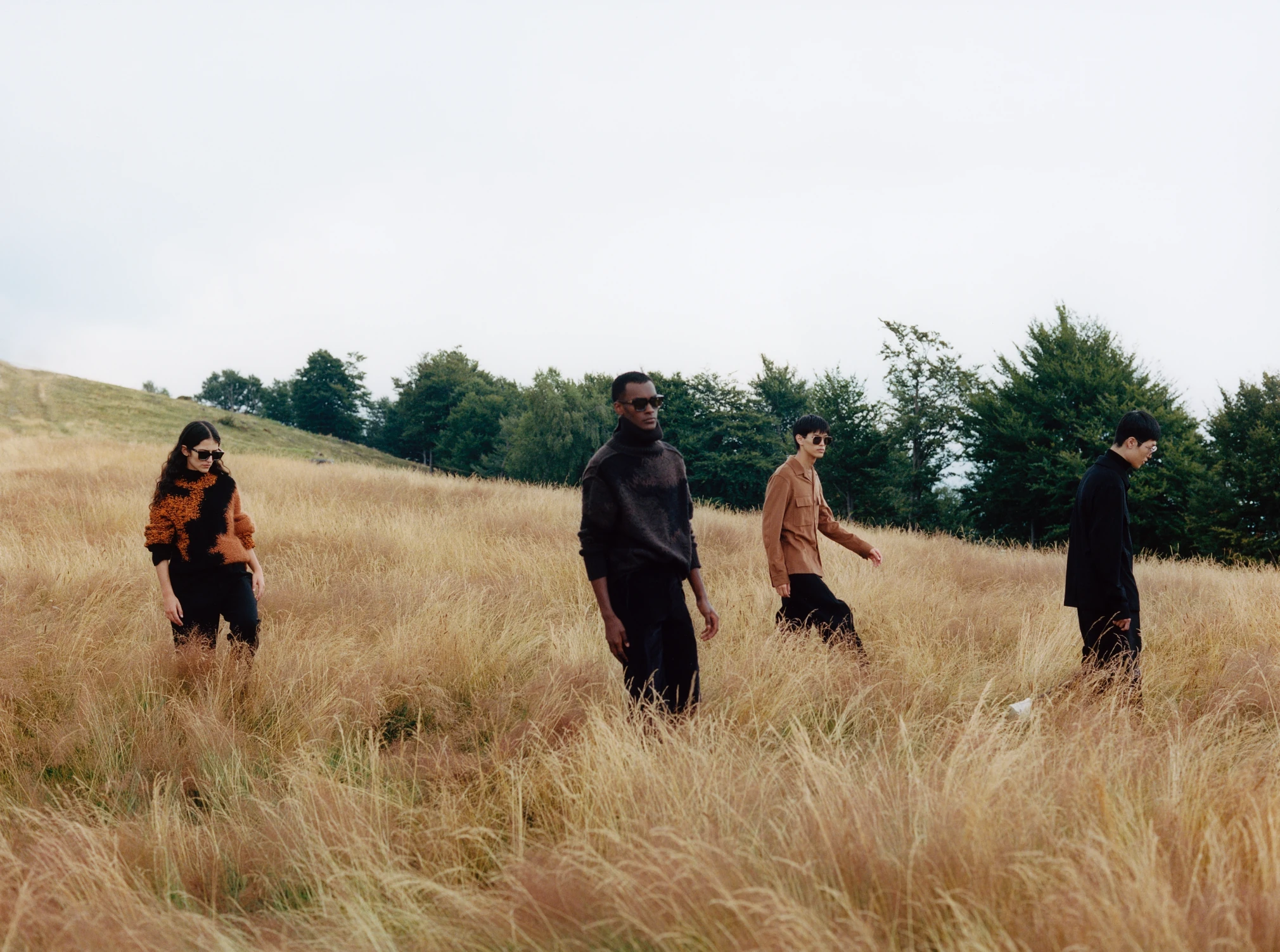 Four models spread across a windblown meadow in black, orange and camel outerwear.